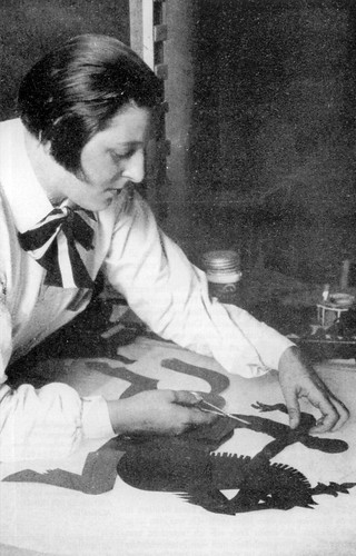 a black and white photo of Lotte Reiniger at her desk working on a shadow puppet