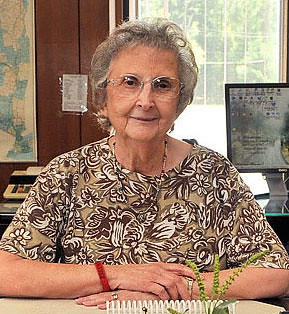 Barbara MacEwen, an elderly white woman white short grey hair and glasses, sits at a desk staring at the camera with a slight smile.