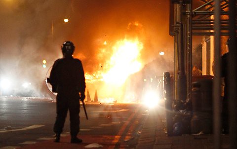 Policeman looks on as vehicles and buildings burn in Tottenham in North London in the midst of a black and working class youth rebellion sparked by the cop killing of a 29-year-old man Mark Duggan. The unrest has spread to other cities. by Pan-African News Wire File Photos