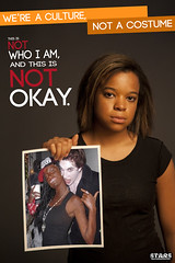 A young, unsmiling black woman holds up a picture of a white woman in black face at a Halloween party. The poster reads We're a culture, not a costume. This is not who I am and it is not okay.