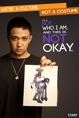 A young, unsmiling Latino man holds up a picture of a costume of a sombrero-wearing white person riding a donkey. The poster reads We're a culture, not a costume. This is not who I am and it is not okay.
