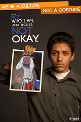 A young, presumably Arab, unsmiling man holds up a picture of a young, jovial white man dressed as a suicide bomber for Halloween. The poster reads We're a culture, not a costume. This is not who I am and it is not okay.