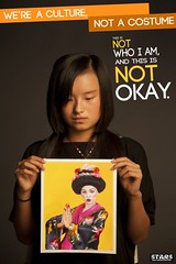 A young, unsmiling, Asian woman looks down at a picture of a white person dressed as a stereotypical Geisha. The poster reads We're a culture, not a costume. This is not who I am and it is not okay.