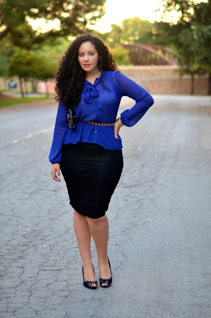 A statuesque, voluptuous woman with dark, curly hair wearing a tight black skirt and silky blue blouse.