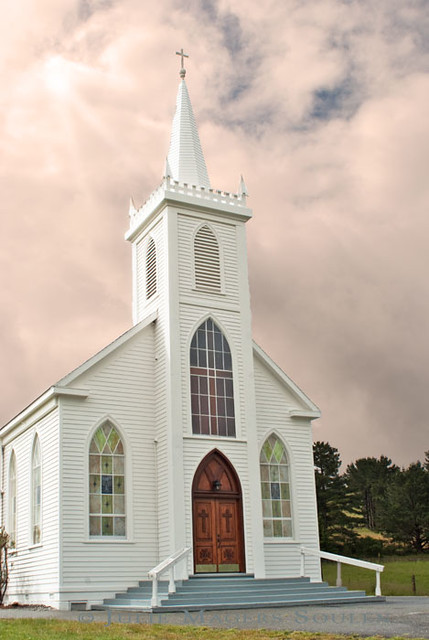 An architectural photo of a white clapboard Christian church with rays  of light shining down from a cloud filled sky.
