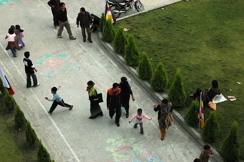 Children play after the initation, walkway from above, Auspicious symbols drawn in chalk, a row of ornamental trees, lawn, Tibetan people, motorcycle, shoes, Buddhist flags, Tharlam Monastery Courtyard, Boudha, Kathmandu, Nepal by Wonderlane