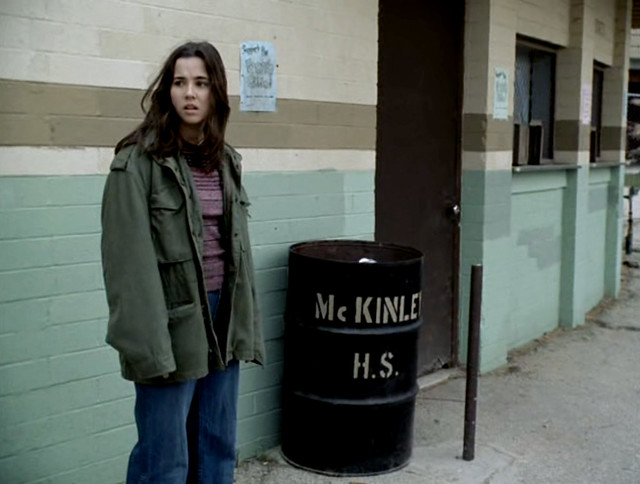 Lindsay Weir, a white teenage girl with dark hair, stands in front of a trash can that says McKinley HS on it