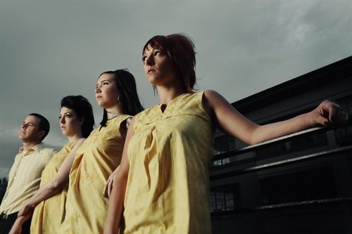 Three women and a man dressed in yellow lean against a car and stare off into the sky.