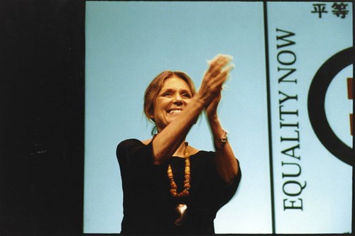 Steinem at a convention; she is standing on stage and clasping her hands