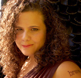Photo of Jaclyn Friedman from the shoulders up. Jaclyn is a white woman with very curly brown hair and a tattoo on her left shoulder that says "brave." She is smiling slightly.