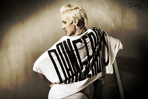 A photograph of Dylan Ryan, a white queer woman with short, bleached blonde hair. She is photographed from the side, wearing a large, flowing shirt that reads DANGEROUS